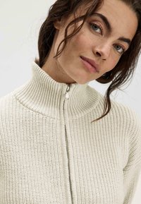 Cremfarbener Reißverschluss-Pullover mit hohem gerippten Kragen, strukturiertem Strickmuster und silbernem Reißverschlussdetails an der Vorderseite.