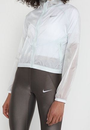 Osoba ubrana w białą siateczkową kurtkę na zamek oraz ciemnobrązowe legginsy Nike z białym logo Nike Swoosh na lewej udzie.