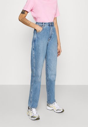 Person trägt hoch taillierte blaue Jeans, ein rosa kurzärmeliges Shirt, weiße und silberne Sneakers und eine Tätowierung am linken Unterarm.