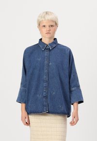 Pārāk liels zilas denim bikses krekls ar apkakli, priekšējiem pogām un sirds formas izgriezumiem. Iespējas plašas piedurknes un brīva piegriezuma.