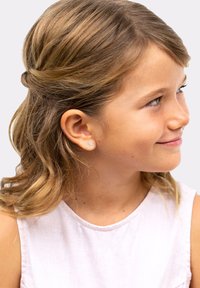 Pequeño pendiente de oro en forma de mariposa, con una superficie texturizada, llevado en la oreja izquierda de un niño con cabello ondulado y castaño claro.
