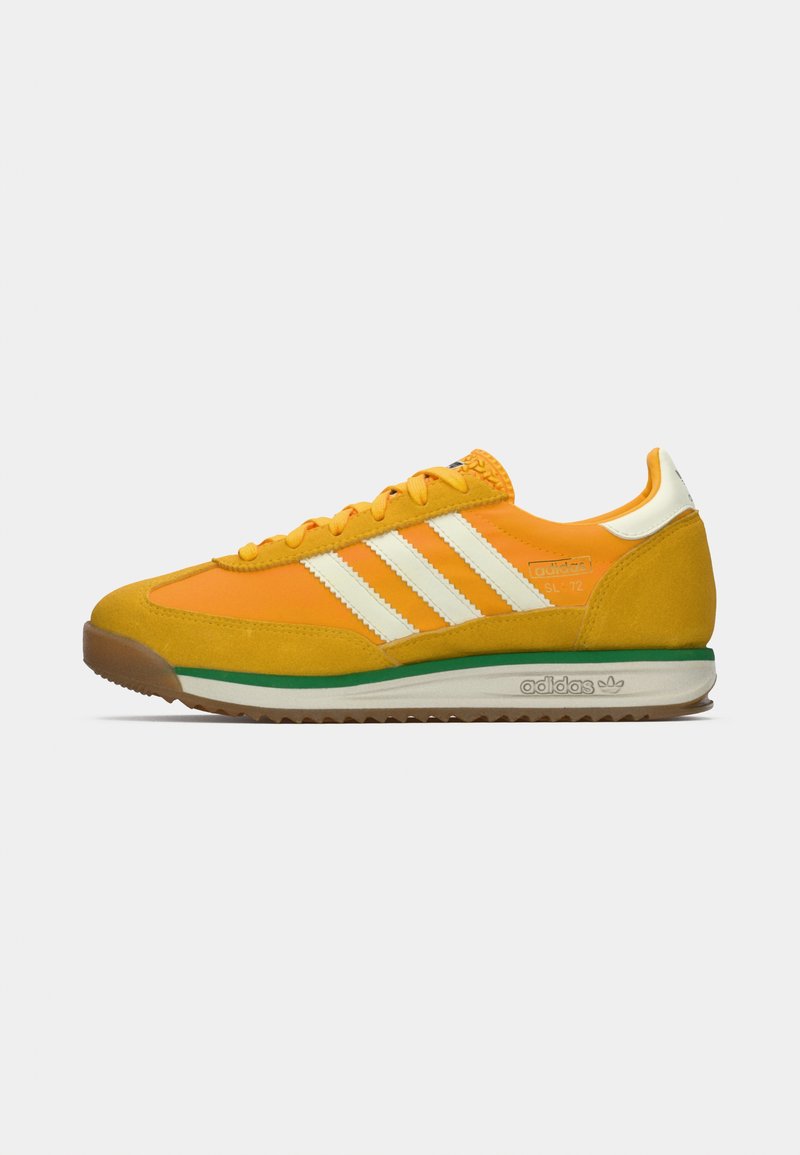 Zapatilla Adidas de perfil bajo en color naranja con superposiciones de ante amarillas, rayas blancas, ribete verde sobre una suela blanca y suela de goma. Vista lateral.