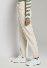 Pantalons en coton beige avec poches latérales et couture visible, associés à des baskets de sport blanches. Le modèle porte un haut rayé vert et blanc.