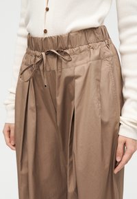 Femme portant un pantalon marron ample à taille haute avec une ceinture élastique et un cordon, associé à un pull côtelé crème avec des boutons.