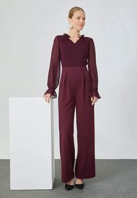 Bordeaux jumpsuit met doorzichtige lange mouwen, met ruches aan de manchetten en een getailleerde top. Hooggewaisted, wijd uitlopende pijpen. Zwarte hakken.