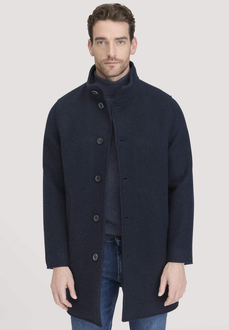 Man in een donkerblauwe knoopjas over een navy coltrui en blauwe jeans, staande tegen een effen lichte achtergrond.