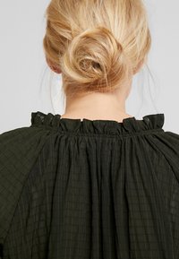Blusa nera con texture, caratterizzata da un colletto arricciato con dettagli di volant, realizzata in un tessuto leggero con sottili motivi a griglia.