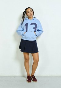 Jasnoniebieska bluza z kapturem z ciemnoniebieskim numerem "13" wydrukowanym z przodu, połączona z granatową plisowaną spódnicą i burgundowymi sneakersami.