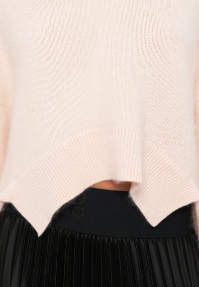 Pull en maille rose clair avec un ourlet côtelé et un design fendu, associé à une jupe plissée noire avec une ceinture texturée.