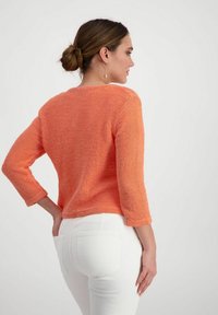 Orange gestrickter Pullover mit Dreiviertelärmeln, Rundhalsausschnitt und strukturiertem Finish, kombiniert mit weißen, eng anliegenden Jeans.