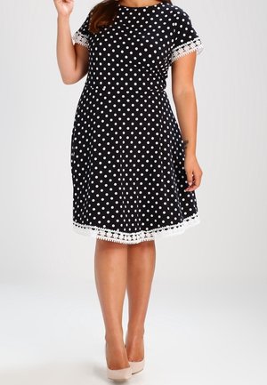 Vrouw draagt een zwarte jurk tot op de knie met witte stippen en kanten randen aan de mouwen en zoom, gecombineerd met beige hoge hakken.