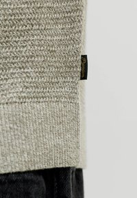 Κοντινό πλάνο από τη μπεζ πλεκτή άκρη πουλόβερ με μαύρη ετικέτα «Premium», φορεμένη πάνω από μαύρο παντελόνι σε απλό φόντο.