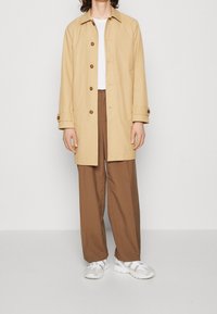 Beige lichtgewicht jas met bruine knopen, witte T-shirt met ronde hals, bruine brede broek en witte sportschoenen. Soepele stof, relaxte pasvorm.