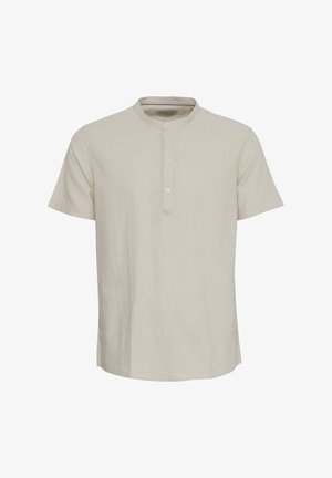 Īsām piedurknēm, gaiši bežs krekls, izgatavots no strukturēta auduma, ar noapaļotu apkakli un puspogu slēgtu ar trim pogām.