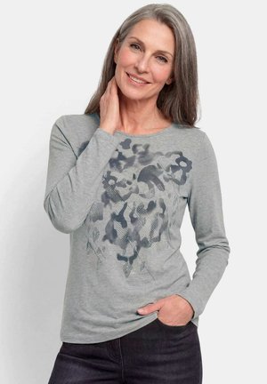 Mittelalte Frau mit grauen Haaren lächelt sanft, trägt ein graues Langarmshirt mit dunklem abstraktem Muster, eine Hand berührt den Hals, die andere in der Tasche.