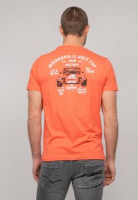 T-shirt en coton orange avec une impression graphique d'une voiture de course et le texte "Indianapolis Race Cup 1989" au dos. Manches courtes, col rond.