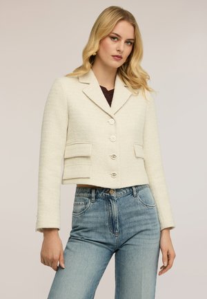 Femme portant un blazer crème texturé avec boutons et un jean taille haute bleu clair, posant devant un fond beige uni.