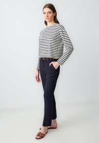 Gestreepte lange mouwen top in crème en marineblauw, gecombineerd met marineblauwe broek. Bruine riem accentueert de taille. Model draagt bruine platte sandalen.