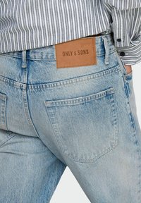 Jean en denim bleu clair avec une apparence délavée, doté d'une étiquette de marque en cuir, de poches arrière et d'une taille standard.