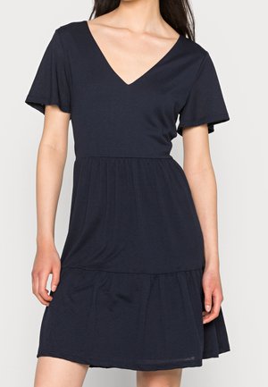 Femme portant une robe courte à manches courtes bleu marine foncé avec un col en V et une taille froncée, debout devant un fond clair uni.