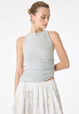 Jeune femme aux cheveux blonds attachés en chignon, portant des boucles d'oreilles argentées en forme d'anneau, un top sans manches gris clair et une jupe plissée blanche avec des motifs floraux subtils.