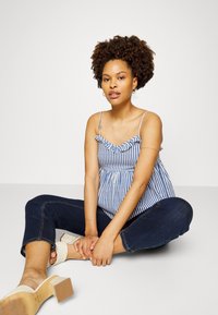 Top sem mangas rayado em azul e branco com decote franzido, combinado com calças de ganga escuras e sandálias de salto bloco bege.