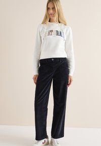 Vit hoodie med "NATURLIG" tryckt i flerfärgade bokstäver, parat med marinblå vida kordbyxor och vita sneakers med röda detaljer.