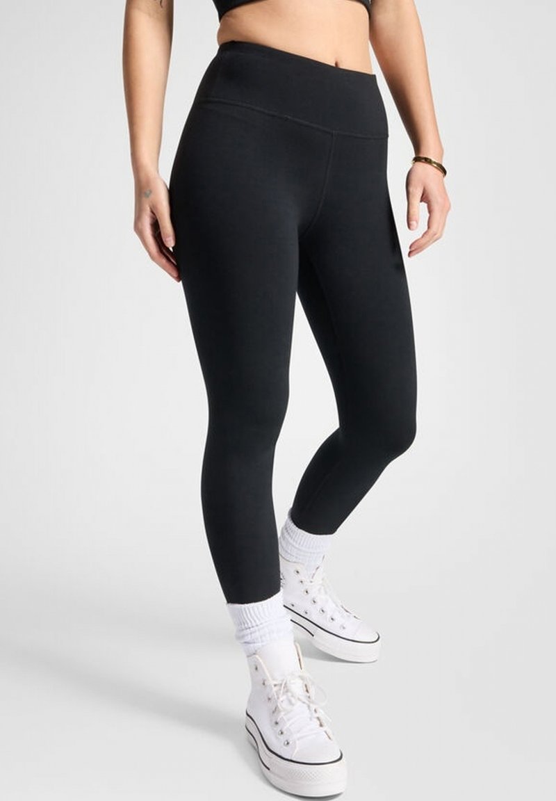Černé legíny vyrobené z elastického materiálu, s vysokým pasem a hladkou texturou, doplněné o bílé vysoké tenisky a ponožky.