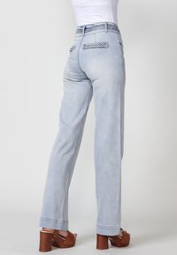 Jeans en denim bleu clair avec une coupe décontractée, des détails tressés au niveau de la taille et deux poches arrière. Accentué par des ourlets larges et des escarpins marron.