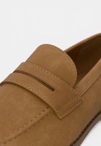 Šviesaus rudo zomšos spalvos bateliai be kulno su apvalia pirštine, dekoratyviniu balnelio dirželiu ir kontrastinės siūlės detale palei pado kraštą. Lygios tekstūros.