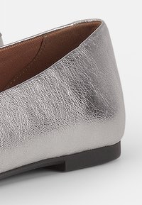 Zilveren platte schoenen met een textuur van metaalachtige afwerking, lage hak en bruine voering. Ronde teen en een glad, glanzend oppervlak.
