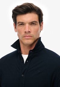 Ciemnoniebieska koszula z kołnierzykiem z detalami w postaci guzików, z teksturowanej tkaniny i wyhaftowanym logo na lewej piersi; noszona na białej koszulce.