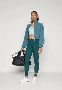 Nő kék sportmelltartóban, türkiz zipzáras dzsekivel, türkiz leggingssel és fehér sportcipővel, fekete Puma tornáz táskával a sima háttér előtt.