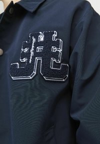 Giacca blu navy con una patch strutturata e un logo distintivo in blu scuro e cuciture bianche; tessuto liscio con design classico a colletto.