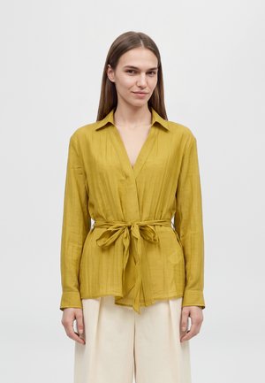 Une femme porte une blouse jaune moutarde ceinturée et un pantalon crème sur un fond clair uni.