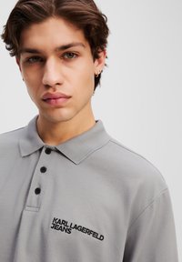 Pelēka polo krekla ar teksturētu apdari, trīs pogām un izšūtu "KARL LAGERFELD JEANS" emblēmu melnā krāsā kreisajā pusē.