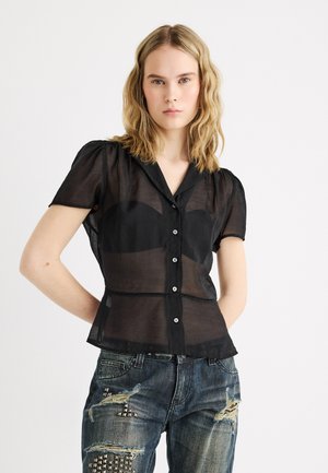 Jeune femme aux cheveux blonds ondulés portant un chemisier noir transparent à manches courtes boutonné et un jean bleu déchiré orné de clous en métal.