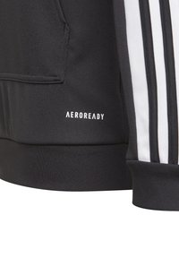 Chaqueta deportiva negra con etiqueta Aeroready, con rayas verticales blancas, textura de tela suave y un bolsillo frontal.