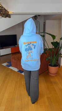 Ljusblå huvtröja med dragsko på huvan, med orange och blå blomgrafik och text på baksidan. Matchas med mörkgrå byxor med vida ben.