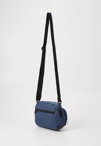 Borsa a tracolla blu in tessuto con superficie testurizzata, dettagli neri, tracolla regolabile e tasca frontale con zip. Design compatto e rettangolare.