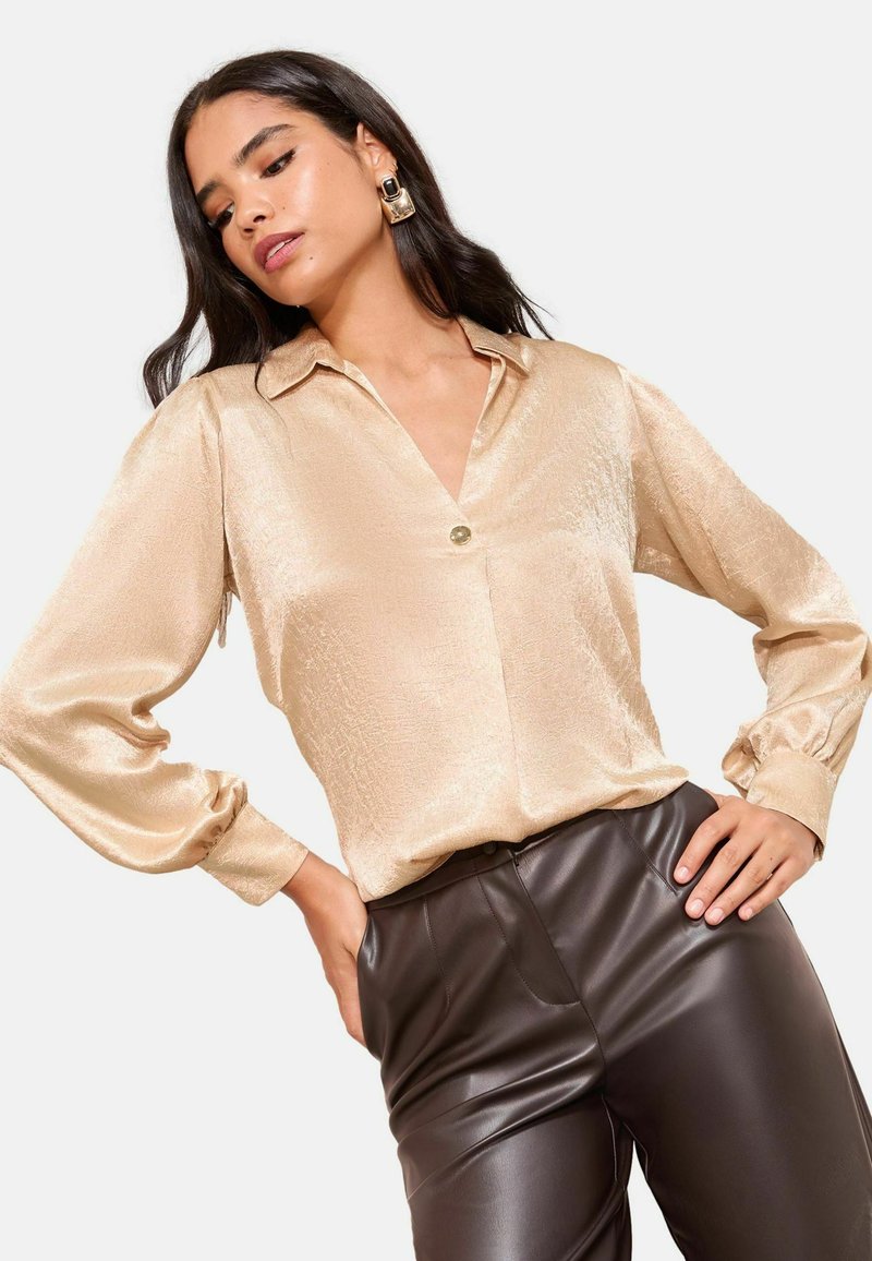 Helles Gold, seidig-bluse mit Kragen, Knopfverschluss und langen Puffärmeln; kombiniert mit braunen Kunstlederhosen.