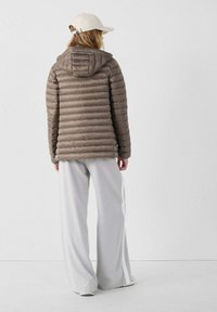 Giacca a vento marrone chiaro con cuciture orizzontali, cappuccio e texture liscia. Abbinata a pantaloni grigio chiaro a gamba larga con strisce laterali bianche.