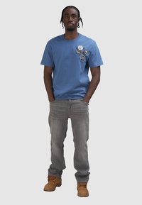 Camiseta de algodón azul claro con parches gráficos en el lado izquierdo del pecho, combinada con jeans grises y botas de trabajo color beige.