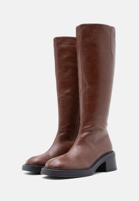 Καφέ δερμάτινες μπότες μέχρι το γόνατο με λεία υφή, στρογγυλεμένη μύτη και χοντρό μαύρο τακούνι. Λεπτομέρεια φερμουάρ στο πλάι.