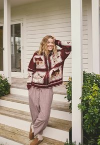 Gestrickte bordeauxrote Strickjacke mit Haus- und Blumenmustern, Reißverschluss; kombiniert mit rot-weiß karierten Hosen und braunen Hausschuhen.