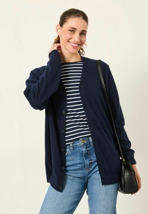 Marineblauer Cardigan mit V-Ausschnitt und Knopfverschluss, kombiniert mit einem marine- und weißgestreiften Oberteil, getragen mit hellblauer Jeans und einer schwarzen Tasche.