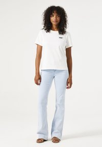 Weißes Baumwoll-T-Shirt mit schwarfer Grafik, kombiniert mit hellblauen Schlagjeans. Model trägt braune Sandalen. Einfarbiger Hintergrund.