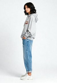 Felpa grigia con tasca frontale e logo ricamato, abbinata a jeans baggy blu chiaro e sneakers bianche. Design semplice, vestibilità rilassata.