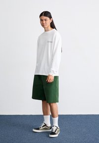 Long-sleeve white cotton shirt with a small black logo, paired with knee-length green cotton shorts. Black and white patterned sneakers complete the outfit.