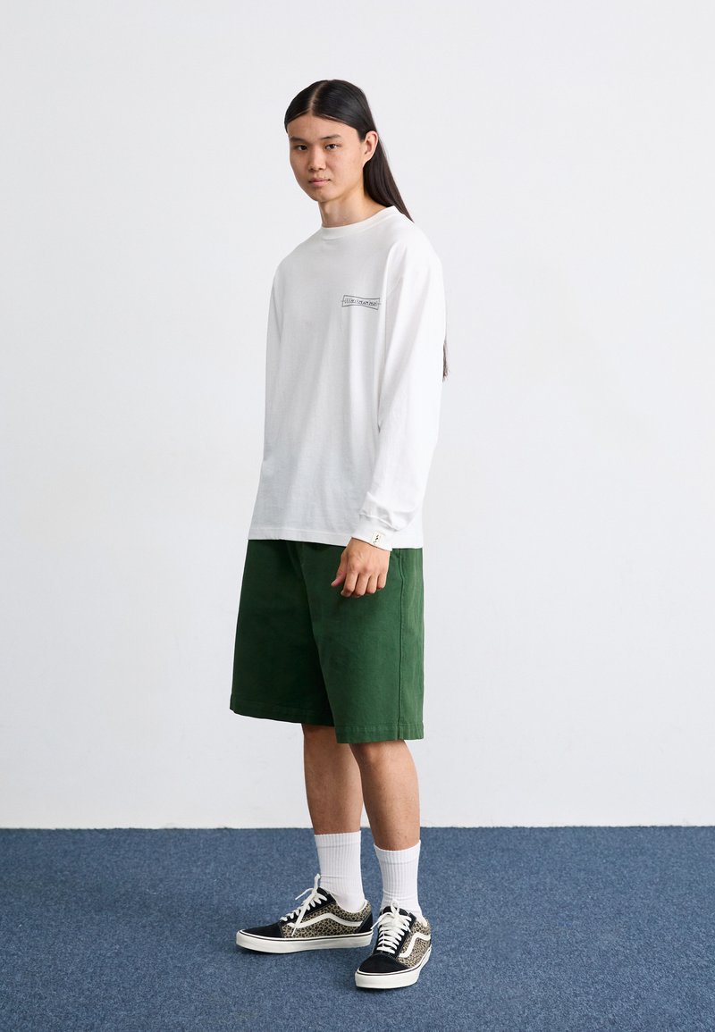 Camisa blanca de algodón de manga larga con un pequeño logo negro, combinada con pantalones cortos verdes de algodón hasta la rodilla. Unos zapatillas de deportes con un patrón en blanco y negro completan el conjunto.
